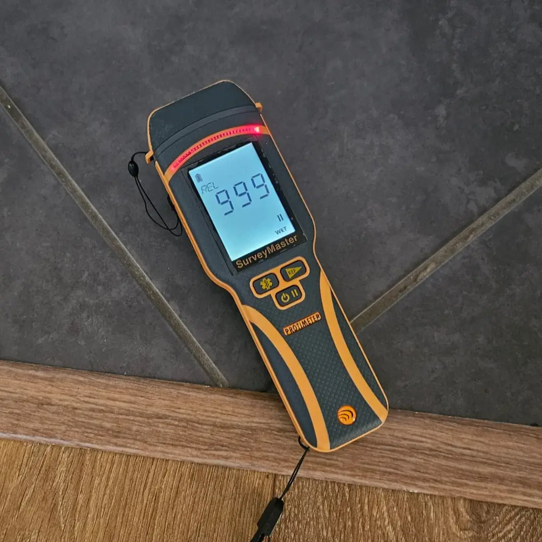 Moisture meter showing 999 WET, indicating significant dampness on a tiled floor.